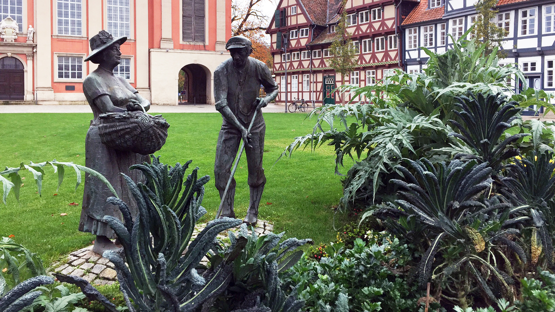 Ein Statue eines Mannes und einer Frau steht auf einer grünen Wiese. Im Vordergrund sind Sträucher angepflanzt. Im Hintergrund stehen mehrere Fachwerkhäuser. Außerdem steht im Hintergrund eine Kirch.