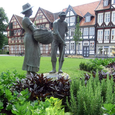 Ein Statue eines Mannes und einer Frau steht auf einer grünen Wiese. Im Vordergrund sind Sträucher angepflanzt. Im Hintergrund stehen mehrere Fachwerkhäuser.