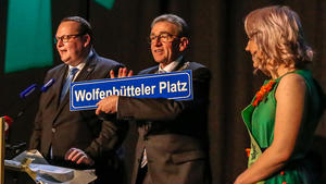 Ein Mann hält ein Schild mit der Aufschrift "Wolfenbütteler Platz" hoch. Neben ihm auf der Bühne stehen ein Mann und eine Frau.