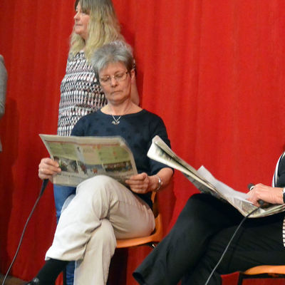 Auf einer kleinen Bühne stehen neben zwei sitzenden Frauen, zwei Frauen. Eine stehende Frau hält in der einen Hand ein Manuskript und der anderen ein Mikrofon, in das sie spricht.