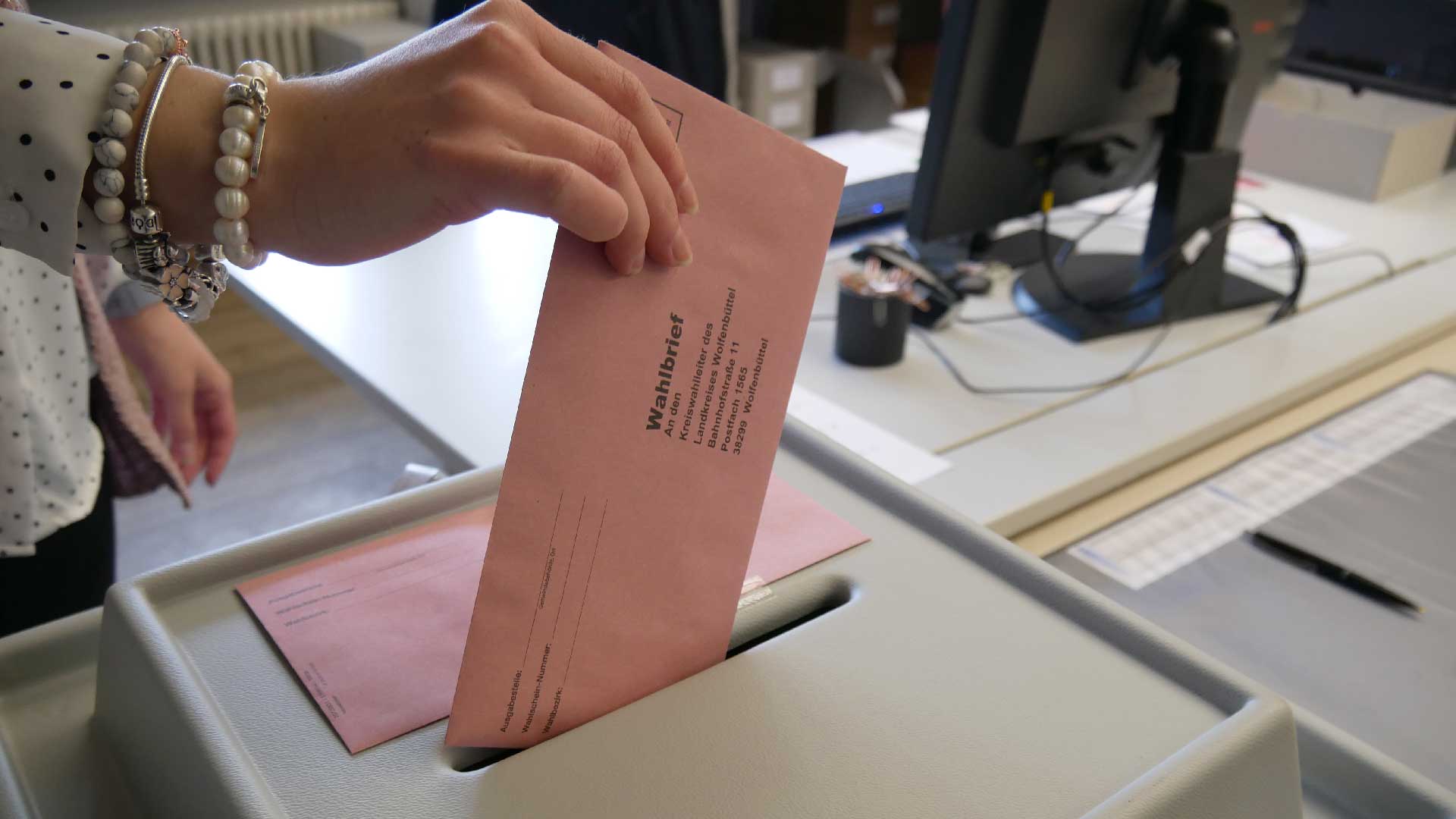 Man sieht die Hand einer jungen Frau, die einen Briefwahl-Umschlag in die Wahlurne einwirft.
