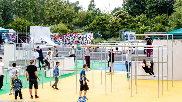 Kinder und Jugendliche spielen auf dem Jugendplatz an Klettergerüsten, im Hintergrund sieht man Beton-Sitzstufen und den Skaterpark.