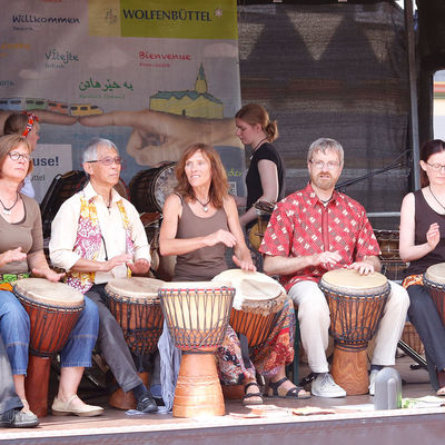 Eine Gruppe von vier Frauen und drei Männer sitzen mit Trommeln auf einer Bühne.