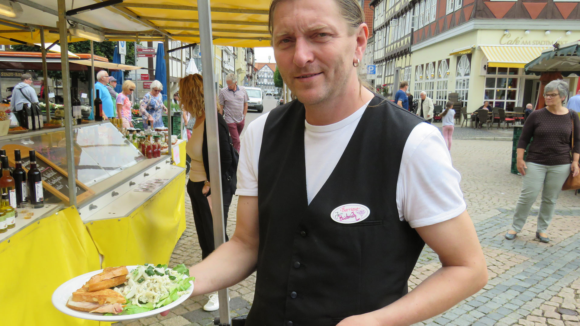 Ein Mann mit einem Teller in der Hand, auf dem zubereitete Speisen liegen, steht vor einem Verkaufsstand auf dem Wochenmarkt in Wolfenbüttel.
