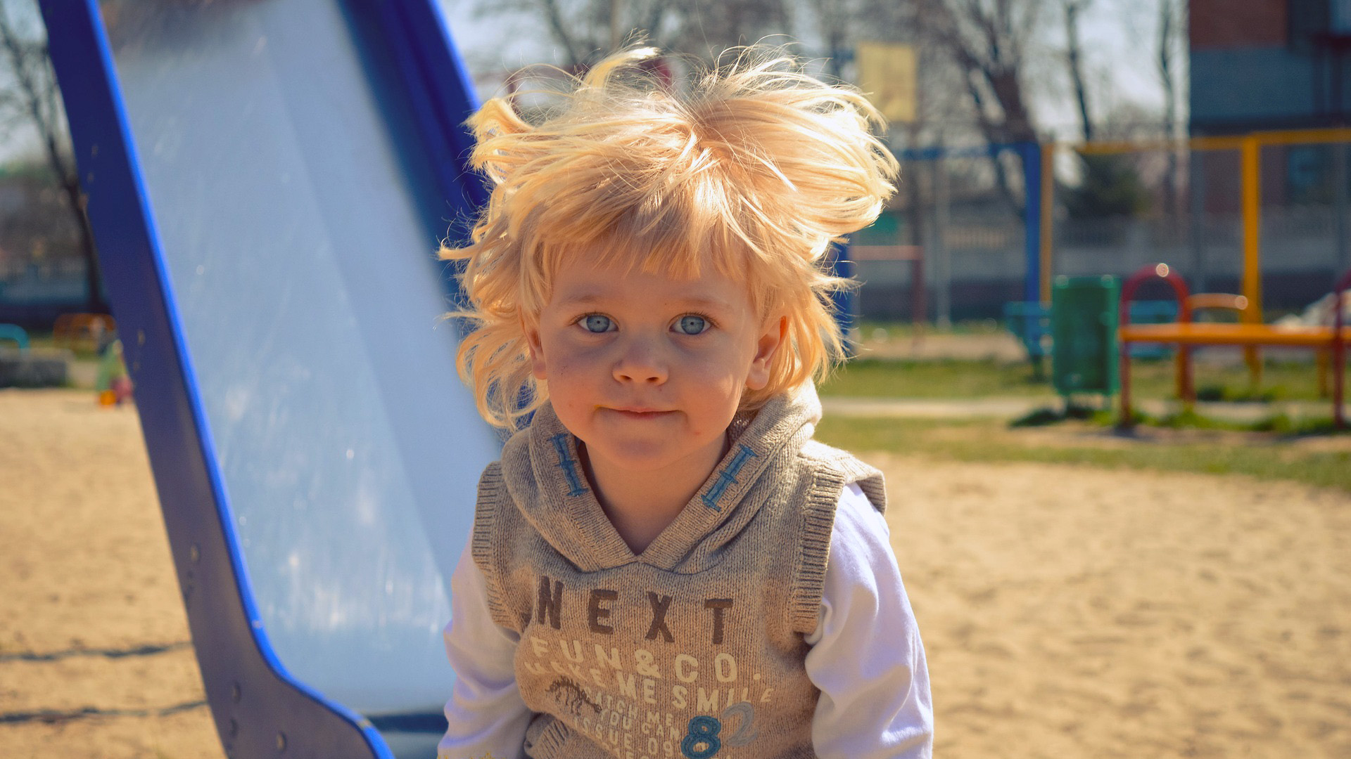 Ein kleiner Junge steht mit wehenden Haaren vor einer Rutsche auf dem Spielplatz und lächelt.