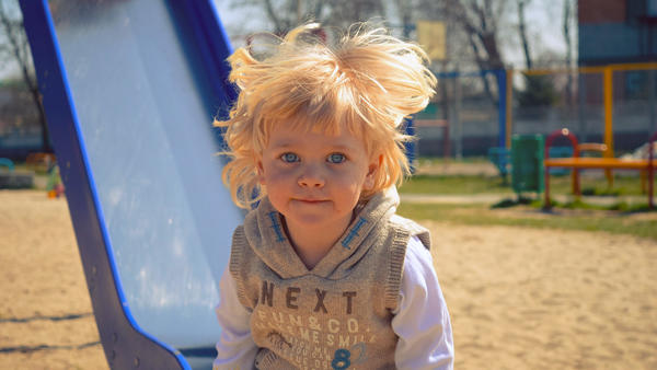 Ein kleiner Junge steht mit wehenden Haaren vor einer Rutsche auf dem Spielplatz und lächelt.