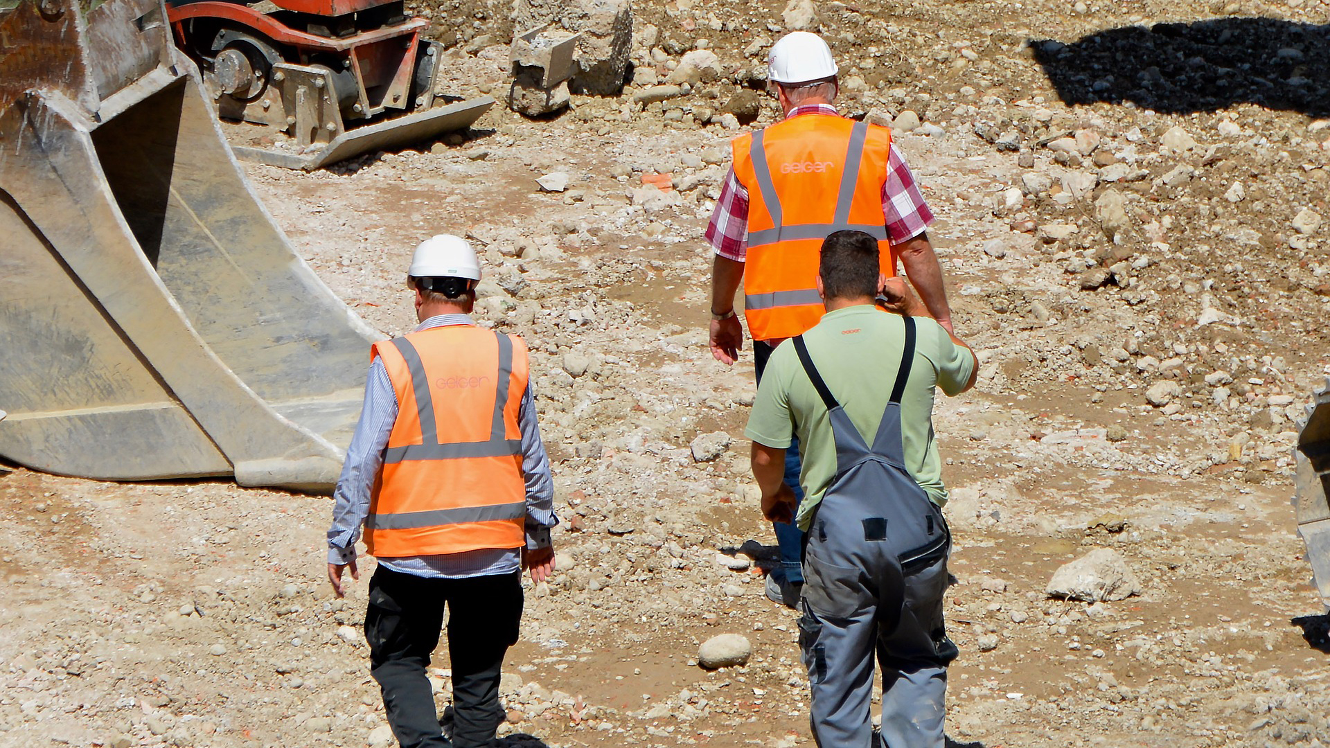 Drei Männer, zwei davon mit Warnweste und Bauhelmen gehen über eine Baustelle.