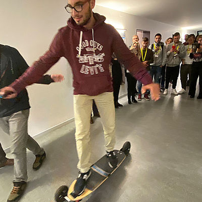 Ein junger Mann fährt auf einem Skateboard einen Flur entlang. Er wird von zwei Männern begleitet, einige Personen schauen dabei zu.