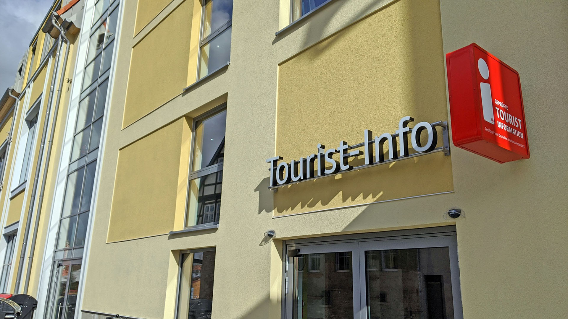 Man sieht eine Hauswand in Gelbtönen, auf der die Leuchtbuchstaben "Tourist-Info" montiert sind. Außerdem hängt daneben eine Leuchtwerbung mit dem offiziellen Zeichen "geprüfte Tourist Information" des Deutschen Tourismusverbandes e.V.