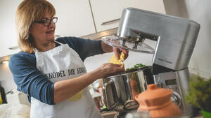 Frau Kücke steht in einer Küche, trägt eine Küchenschürze und hantiert mit Kartoffelteil an einer Küchenmaschine.