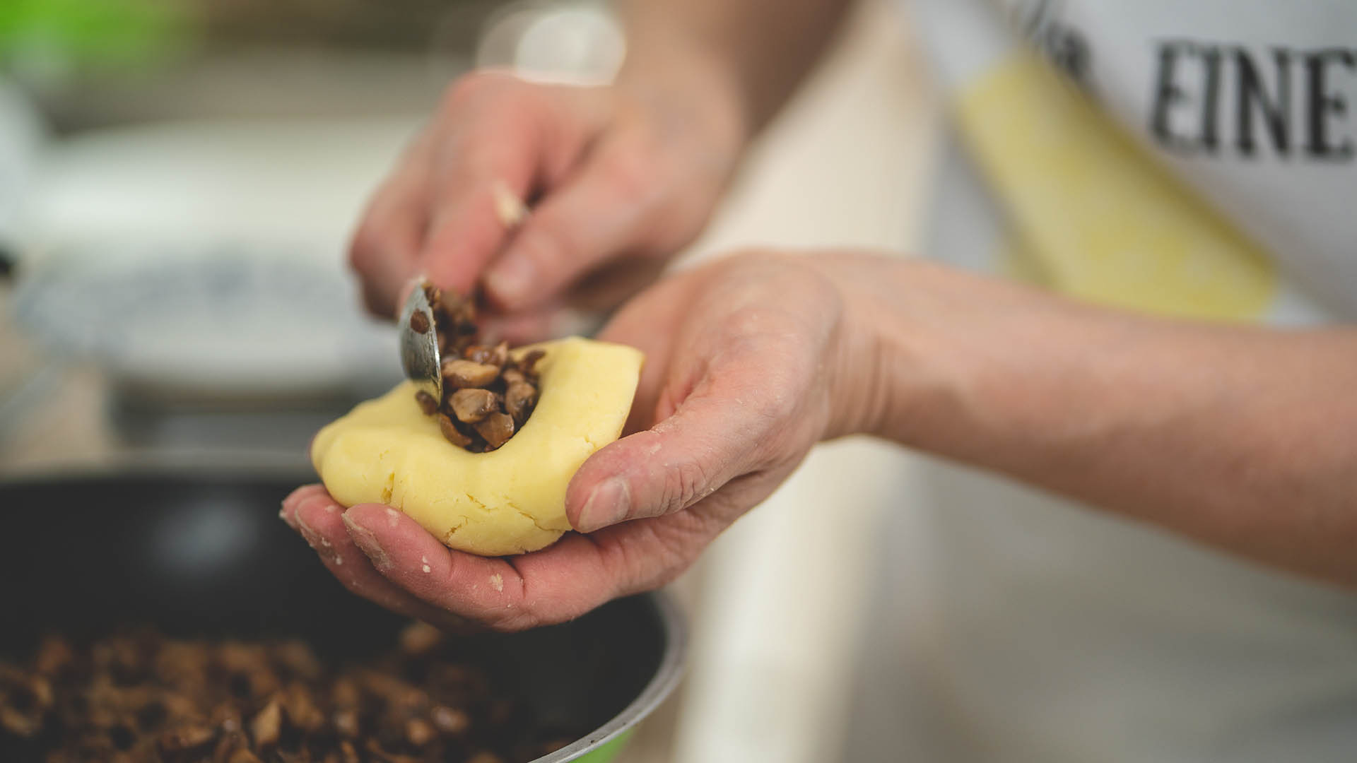 Eine Frau hält ein Stück Kartoffelteig in der Hand und füllt mit der anderen Hand mt dem Löffel kleingeschnittene Pilze in den Teig.