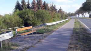 Am Rand eines Fahrradweges steht eine Sitzbank. Am rechten Bildrand ist die anschließende Landstraße zu erkennen.