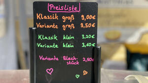 Auf einem schwarzen Preisaufsteller sind in verschiedenen Neonfarben fünf verschiede Cheesecake-Varianten und zugehörige Preise ausgewiesen.