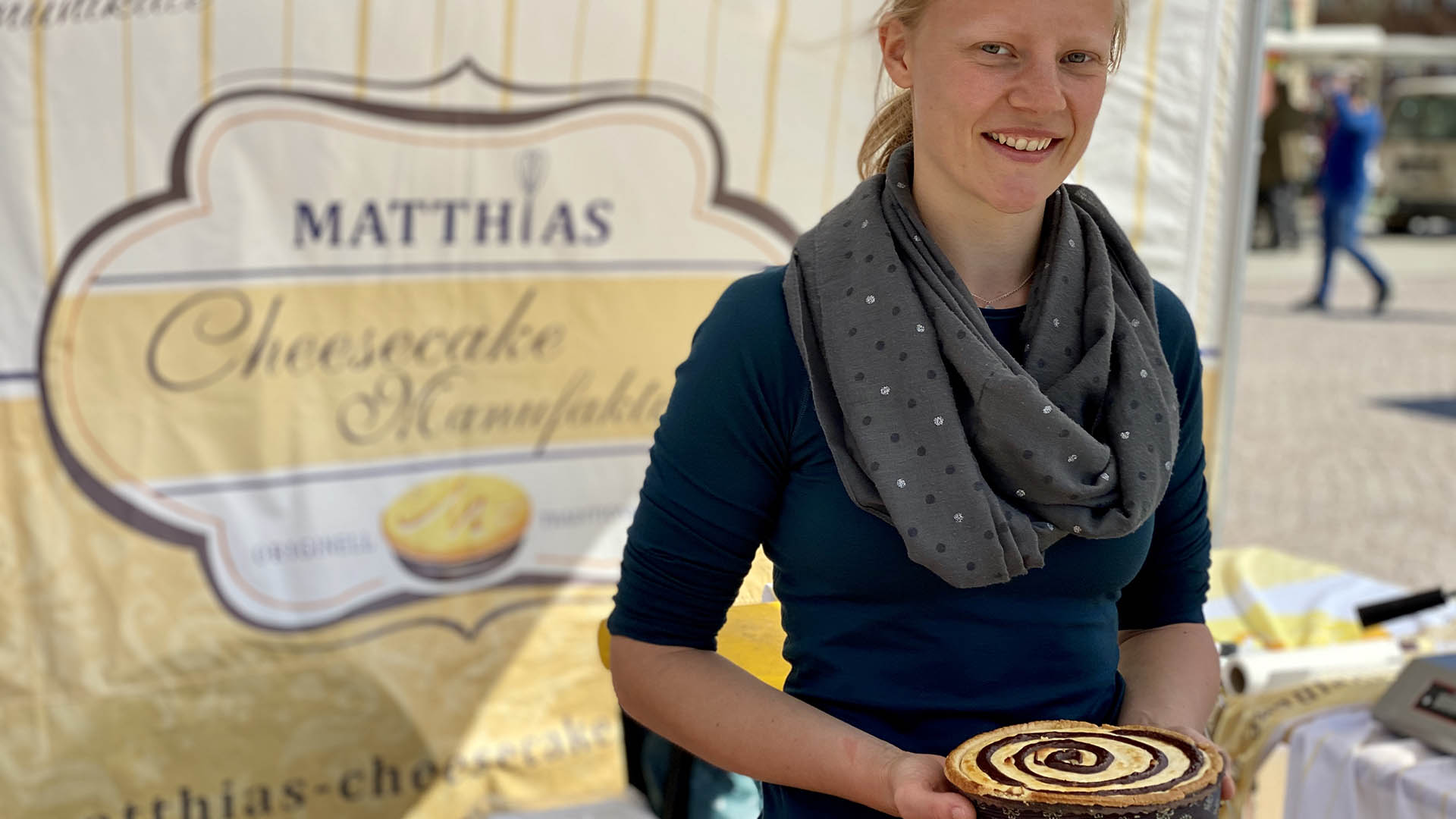 Eine junge Frau hält einen Käsekuchen in den Händen vor sich und lächelt in die Kameria. Hinter ihr hängt ein Werbeplakat von "Matthias Cheesecake Manufaktur".