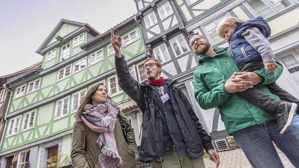 Stadtführer Sebastian Mönnich steht mit einer jungen Familie - Mann, Frau und Kind- vor einer Reihe von Fachwerkhäusern und weist mit ausgestrecktem Arm auf eine Sehenswürdigkeit.