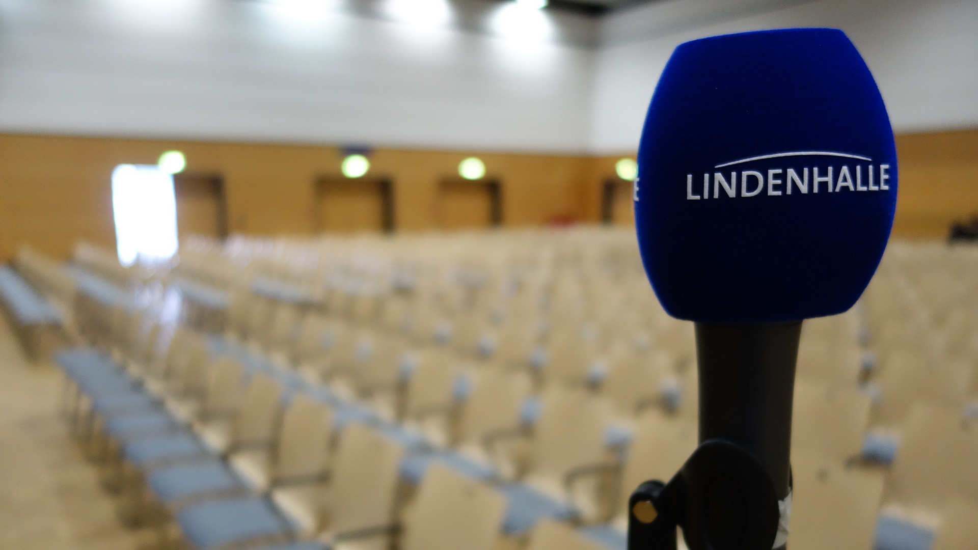Eine blaues Mikrofon mit der Aufschrift "Lindenhalle". Im Hintergrund sind schemenhaft viele Stuhlreihen zu erkennen.