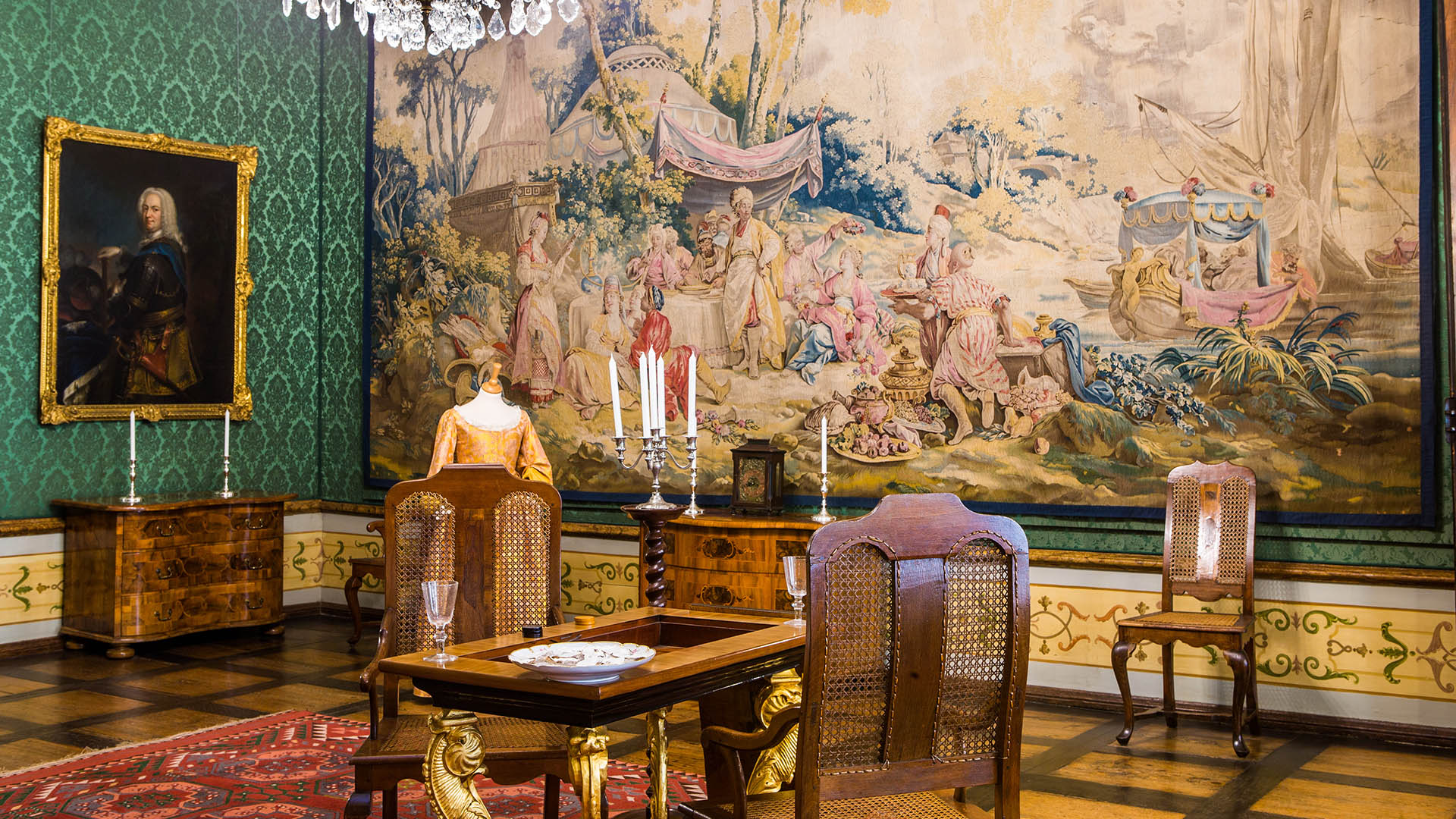 Das Foto zeigt eines der Vorzimmer der Wolfenbütteler Herzogin mit einem roten Teppich auf edlem Holzfußboden und einem Backgammon-Spieltisch. An der Wand befindet sich ein großer Wandteppich einer französischen Manufaktur, auf dem viele orientalisch gekleidete Personen vor einem Zelt speisen.