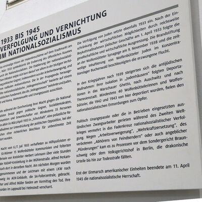 Das Foto zeigt eine große weiße Wandplatte. Der in schwarz aufgedruckte Text beschreibt die Verfolgung der politischen Gegner des NS-Regimes und der Wolfenbütteler Juden.