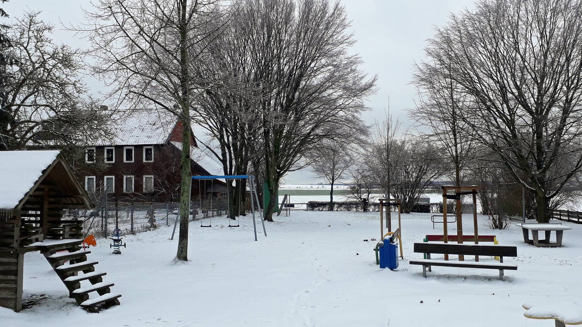 Blick über den verschneiten Spielplatz mit verschiedenen Spielgeräten. Im Hintergrund stehen Bäume und ein Haus.