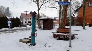 Blick auf einen Dorfplatz mit einer Bank, Bushaltestelle und einer alten Wasserpumpe. An der Pumpe und an einem Straßenschild sind Meisenknödel befestigt.
