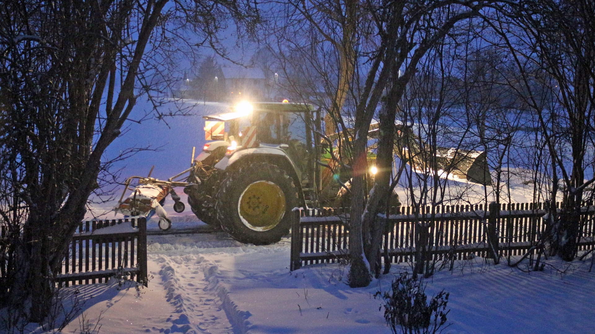 Vor einem Gartentor fährt in der Dämmerung ein beleuchteter Traktor durch den Schnee.