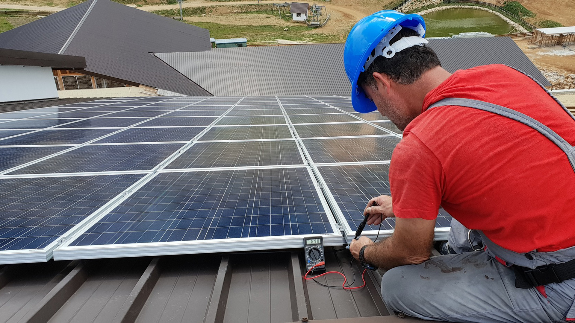 Ein Mann in Arbeitskleidung und Sturzhelm sitzt auf einem Dach und montiert Photovoltaik-Platten.