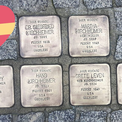 In einem Kopfsteinpflaster sind sechs Platten aus Metall eingelassen mit Namen von sechs Personen, ihren Geburtsdaten und dem Hinweis, dass sie in die USA geflohen sind und den Holocaust überlebt haben. Daneben liegt ein rotes Herz aus Papier.