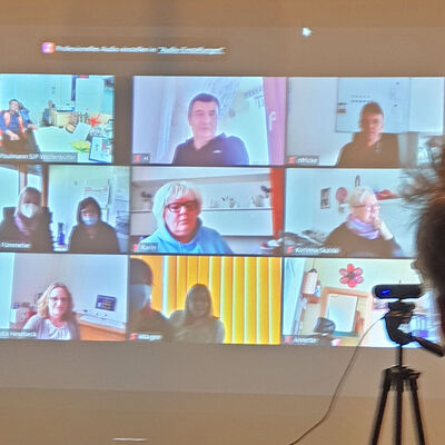 Auf einer Leinwand sind zwölf Teilnehmerinnen und Teilnehmer in einer Videokonferenz zu sehen, ein Mann sitzt vor der Leinwand.