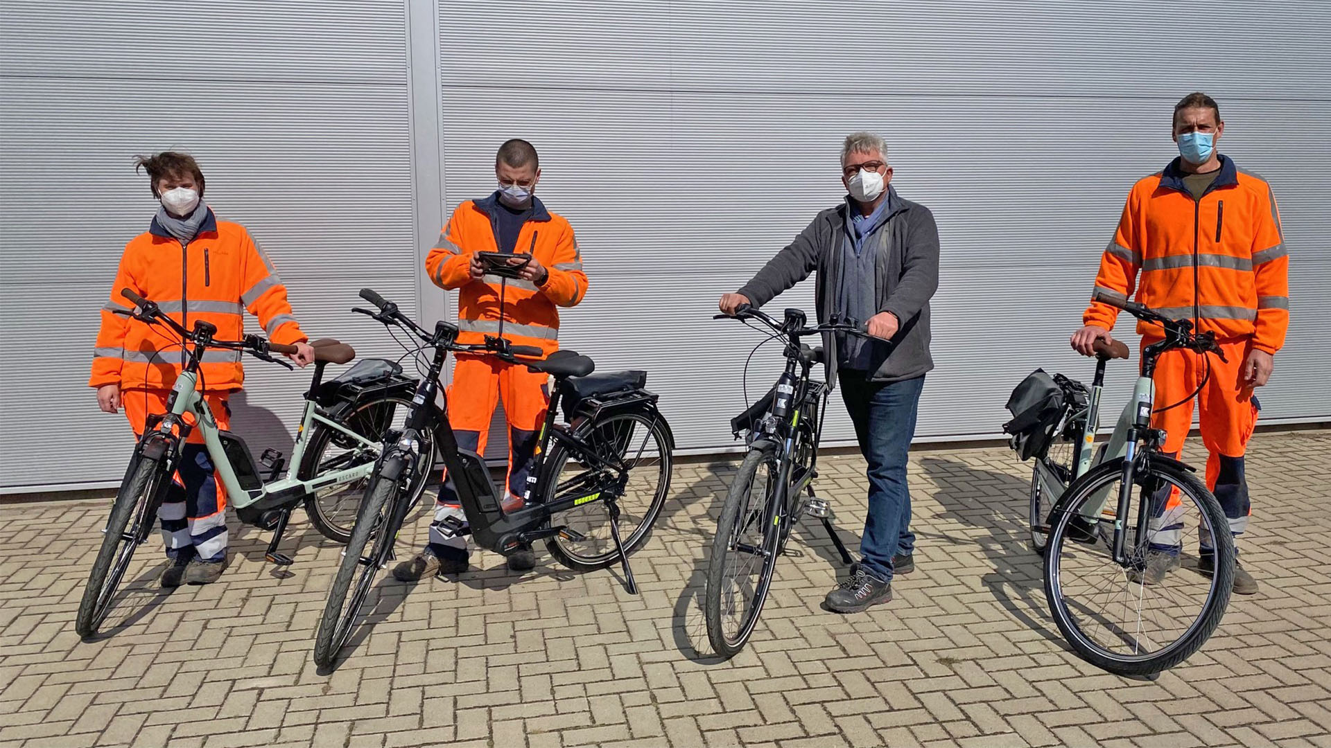 Vier Männer, zum Teil in oranger Dienstkleidung, stehen neben je einem E-Bike.