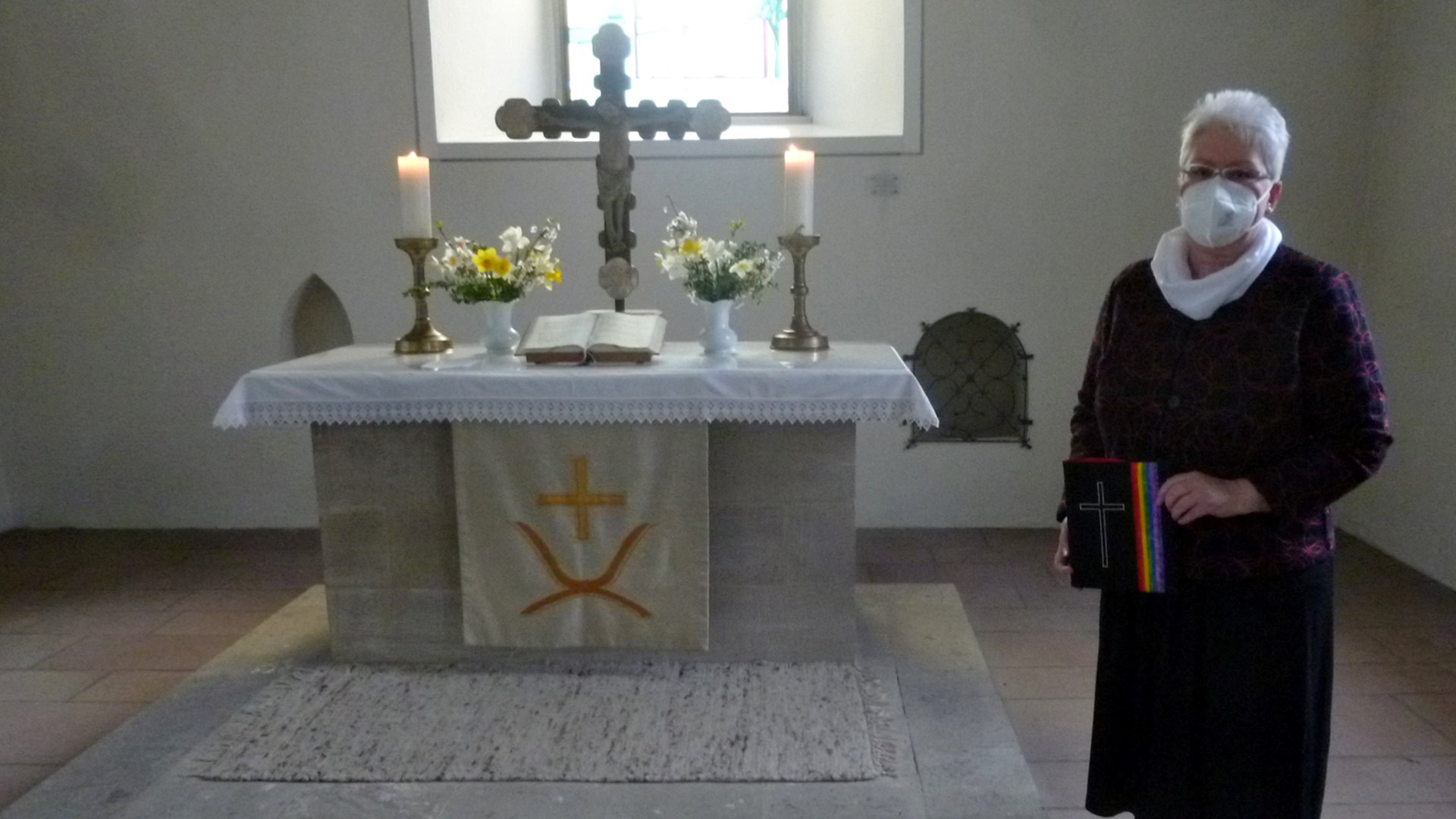 Vor einem Altar mit Blumensträußen, einem großen Kreuz, einem aufgeschlagenem Buch und zwei Kerzen steht eine Frau in einem dunklen Kleid und einer Mund-Nase-Maske.