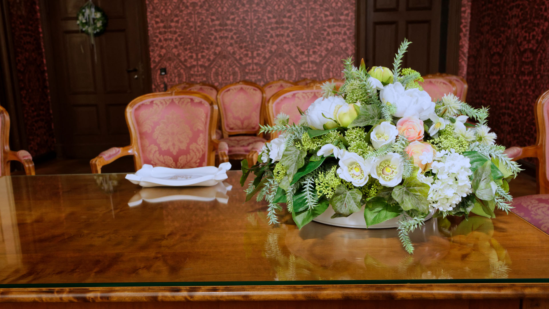 Blick in einen Raum mit Parkett, historischen mit Schnitzereien verzierten Schränken, rötliche Tapeten mit Prägung. Im Vordergrund steht ein Tisch mit Blumengesteck, dahinter sind Holzstühle mit rosa Stoffbezügen aufgestellt.