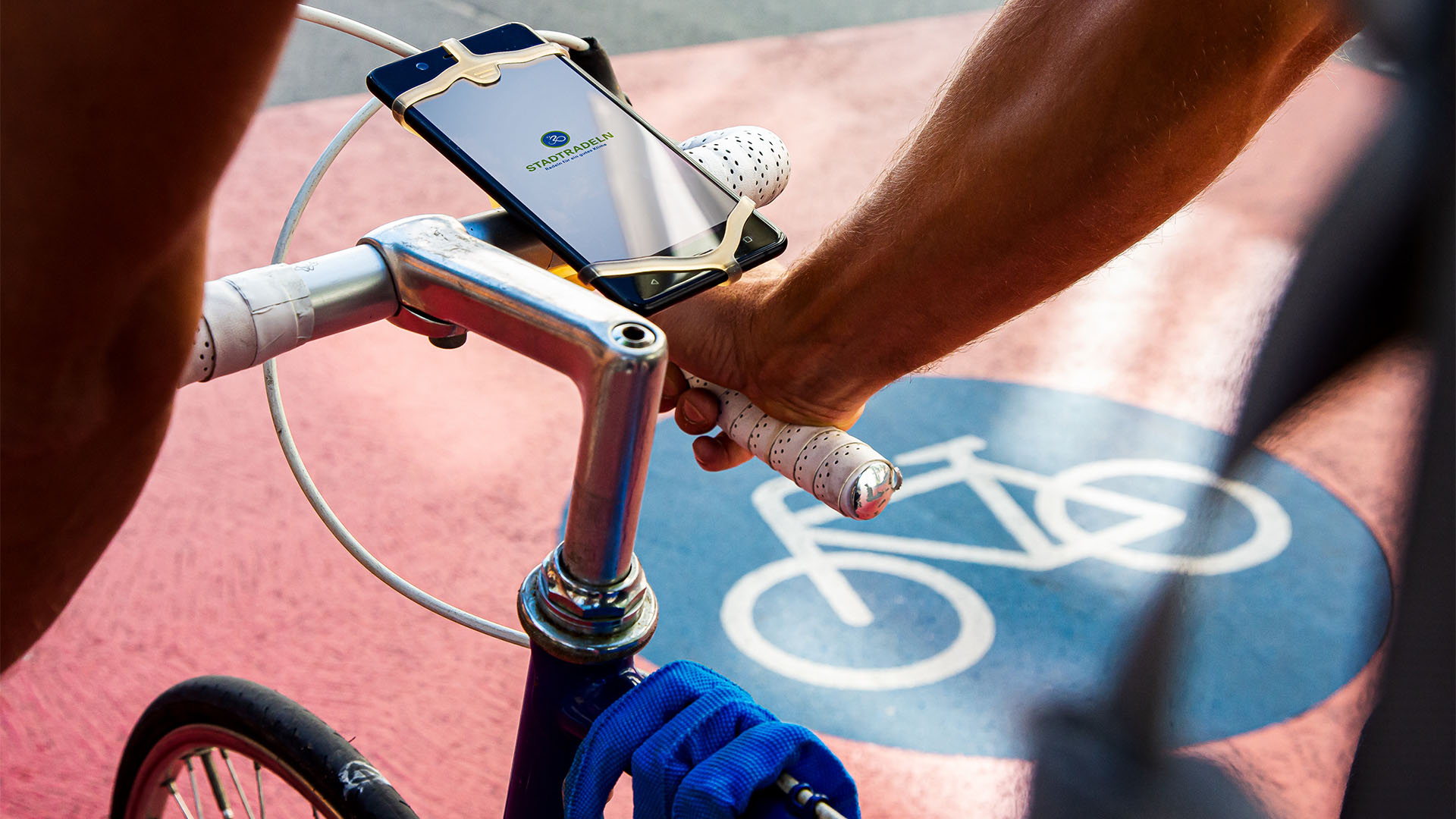 Sicht auf einen Fahrradlenker an dem ein Handy angebracht ist. Im Hintergrund ist auf dem Boden das Verkehrszeichen für Radweg aufgebracht.
