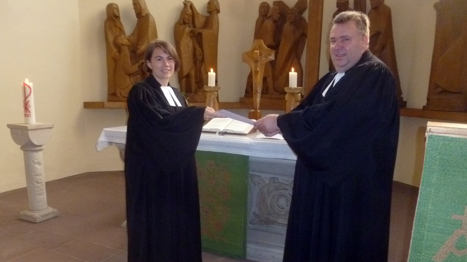 Propst Dieter Schultz-Seitz überreicht Pfarrerin Julia Jansen eine Urkunde. Dahinter steht ein Altar. Hinter dem Altar stehen mehrere Holzfiguren.