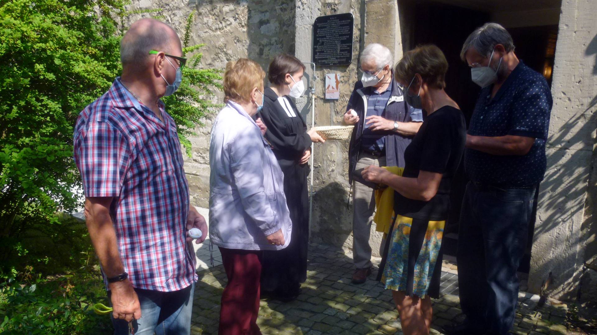 Vor dem Eingang zur Kirche stehen sechs Personen. Die Pfarrerin hält ein Körbchen in der Hand aus dem sich die anderen Personen kleine Cremetuben nehmen.