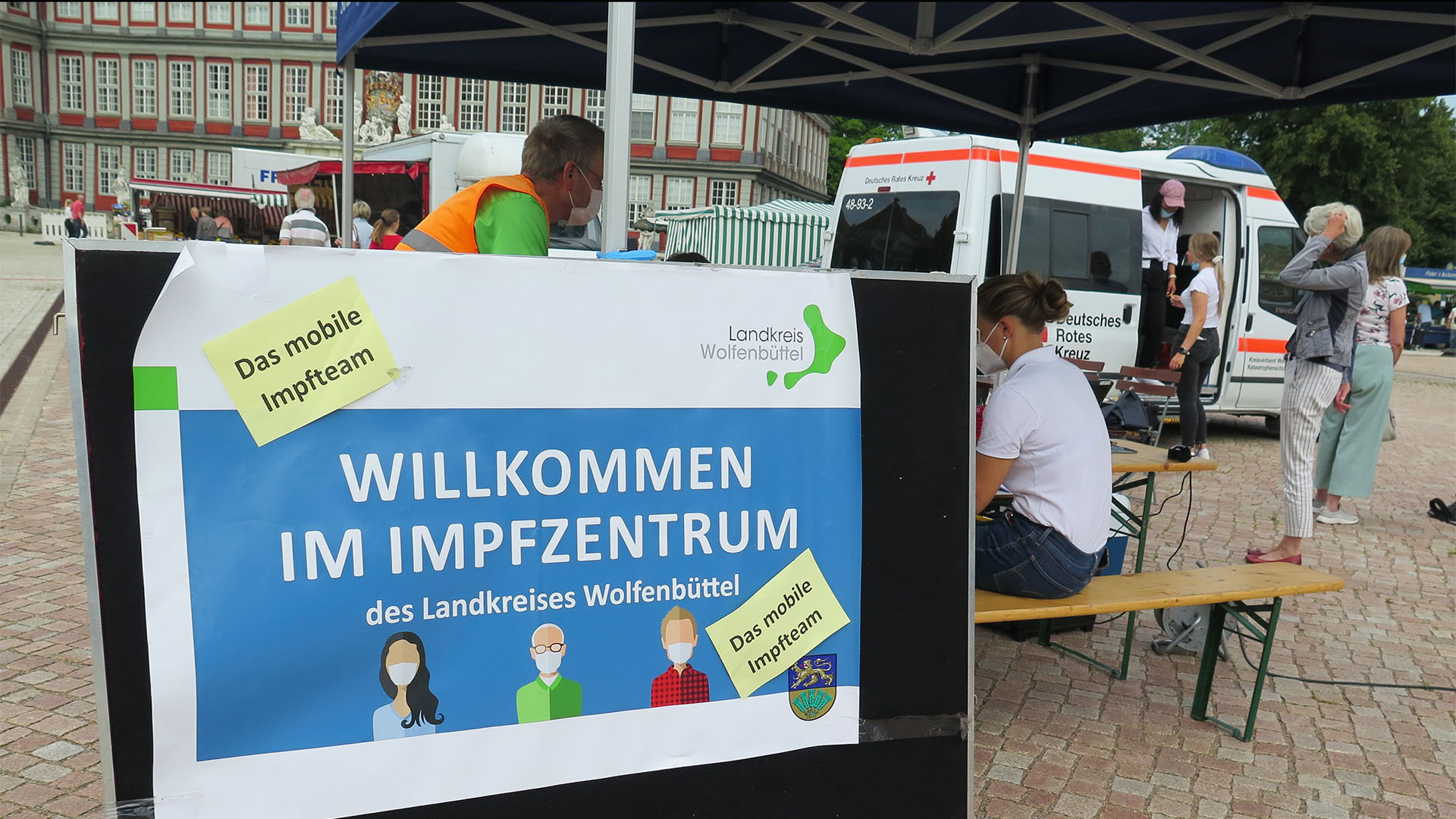 Der Impfbus steht auf dem Schlossplatz, im Hintergrund das Schloss. Im Vordergrund sitzen DRK-Mitarbeiter hinter einem Plakat "Willkommen im Impfzentrum des Landkreises Wolfenbüttel - das mobile Impfteam".