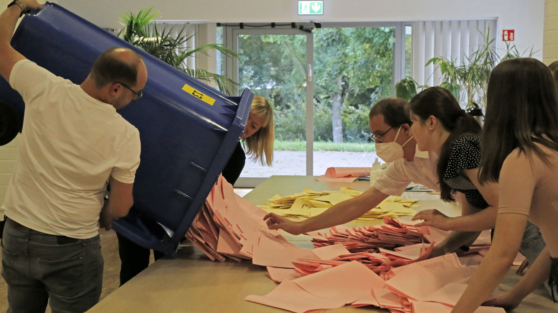 Zwei Personen schütten die Stimmzettel aus einer Wahlurne auf einen Tisch, weitere Personen auf der anderen Tischseite passen auf, dass nichts vom Tisch fällt.