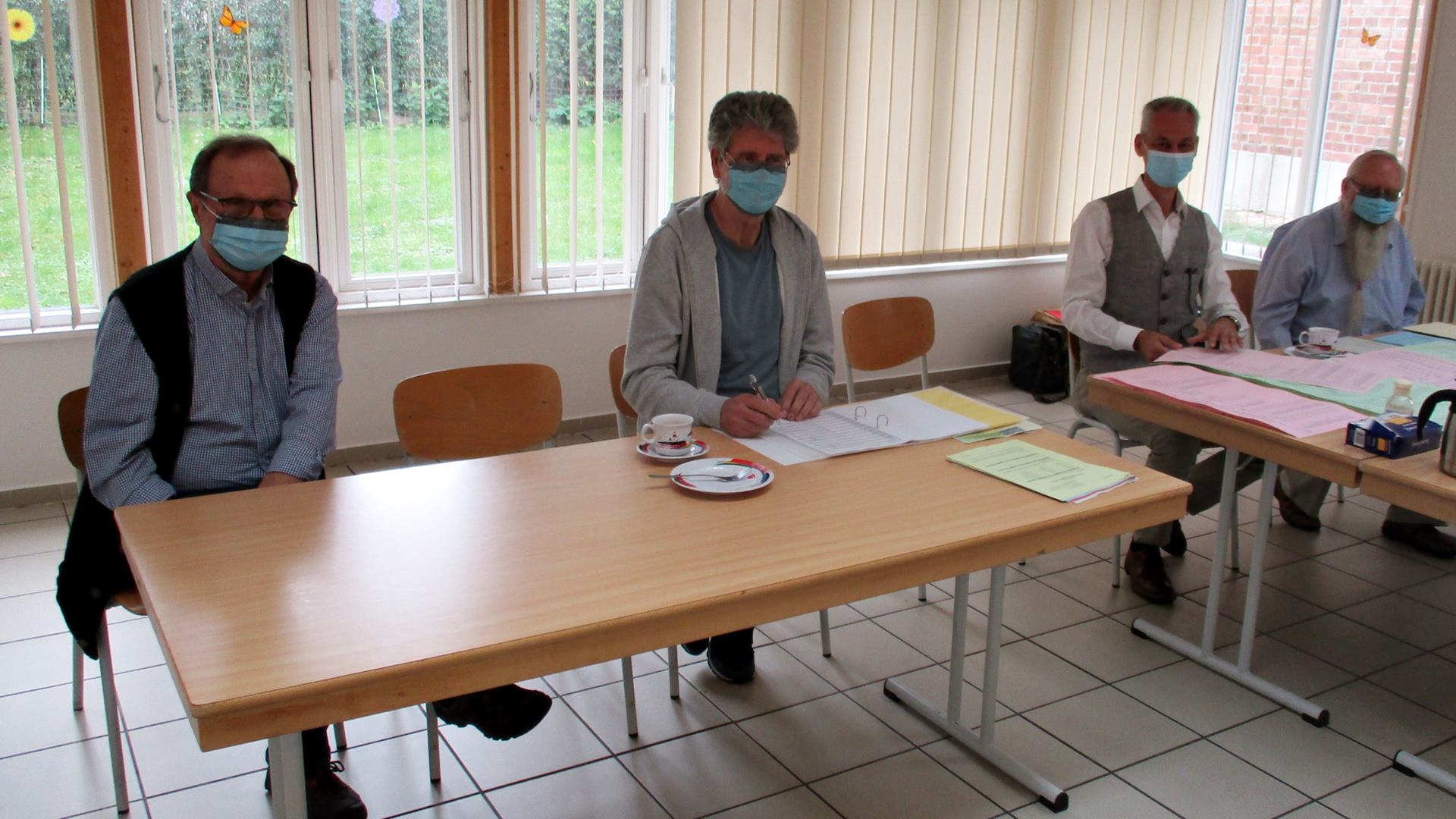 Vier Personen mit blauem Mund-Nase-Schutz sitzen an Tischen mit Wahlunterlagen und Stimmzetteln.