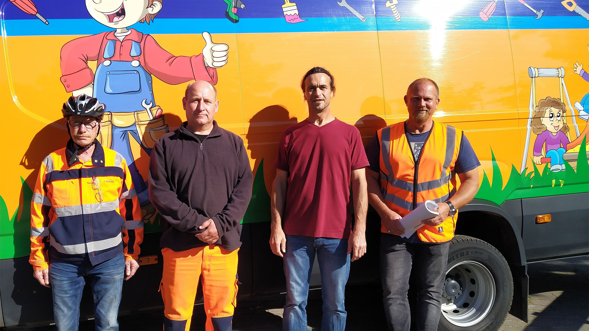 Vier Männer, teilweise in Arbeitskleidung, stehen vor einem großen orangen Fahrzeug mit Comicfiguren.