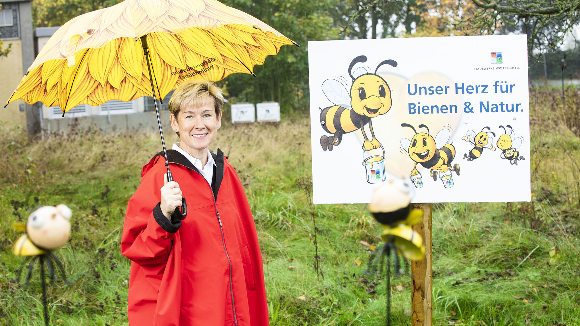 Eine Frau mit Regenschirm steht vor einer Wiese und einem Schild "Unser Herz für Bienen & Natur".