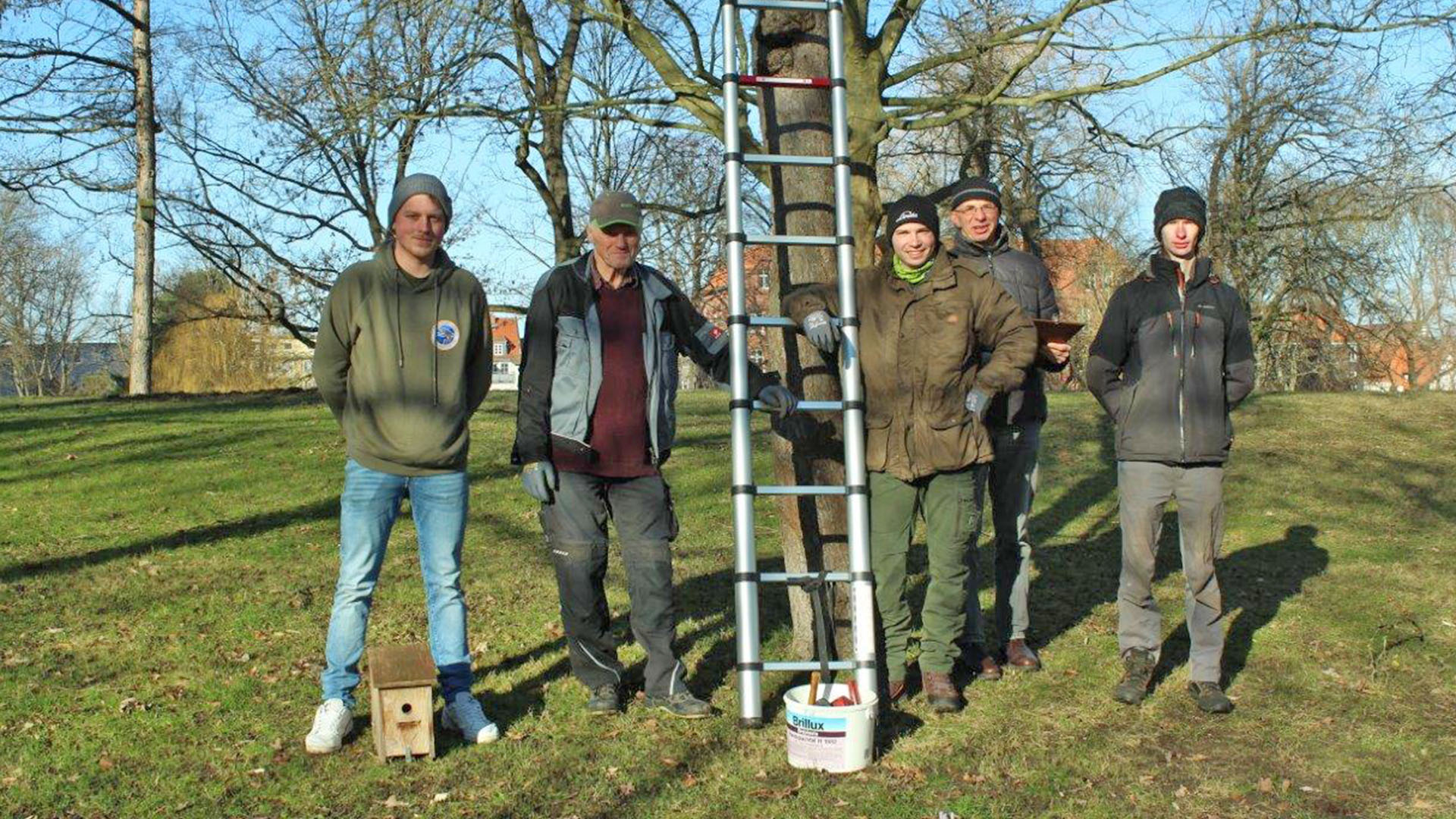 Fünf Männer stehen neben einer Leiter, die an einen Baum gelehnt ist, auf dem Boden steht ein Nistkasten.