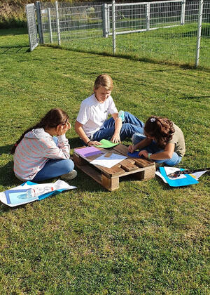 Eine Frau und zwei Mädchen sitzen auf einer Rasenfläche und basteln oder schreiben.