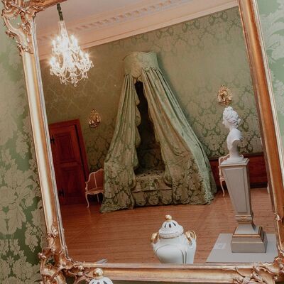 Blick in einen Wandspiegel, darin sieht man das Geburtszimmer der Herzogin Anna Amalia mit einem Himmelbet, einer Büste, Kronleuchter und kleinen Dekogegenständen