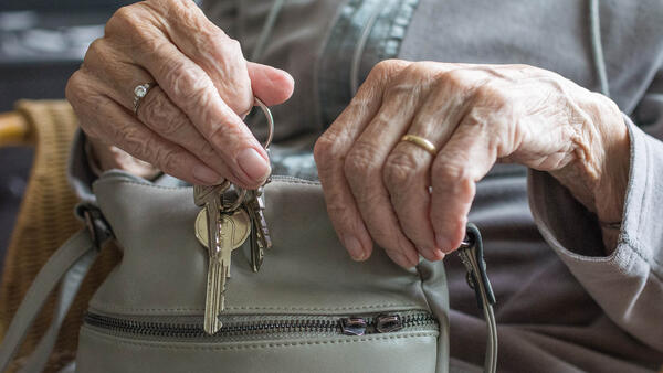 Eine alte Frau hält eine Handtasche auf dem Schoß und einen Schlüsselbund in einer Hand.
