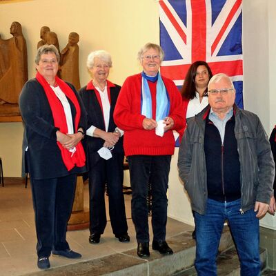 Fünf Personen lächeln in die Kamera. An der Wand ist eine britische Flagge zu erkennen. Die Personen stellen das Kirchen-Team dar.