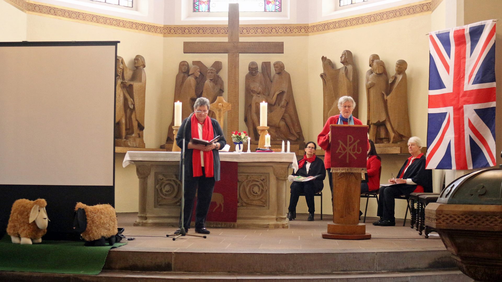 Vor einem Altar stehen zwei Frauen, die eine Lesung halten. An der Wand ist eine britische Flagge zu erkennen. Im Hintergrund sitzen drei Frauen und hören zu.