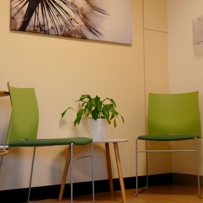 Ein kleiner Tisch in einer Raumecke zusammen mit zwei grün bezogenen Stühlen . Auf dem Tisch steht eine Grünpflanze. Rechts und links stehen kleine Baumstämme.