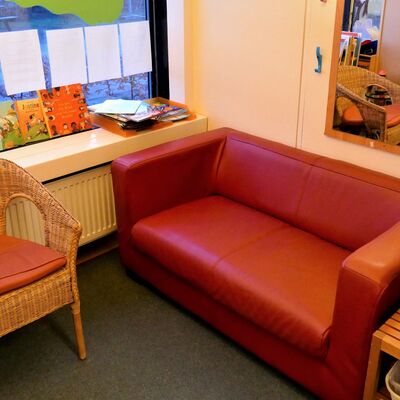 Eine Sitzecke mit einem roten Sofa, einem Korbsessel mit rotem Kissen einem kleinen Holztisch und ein großer Wandspiegel. Am Fenster im Hintergrund hängen Zettel und stehen Bücher.