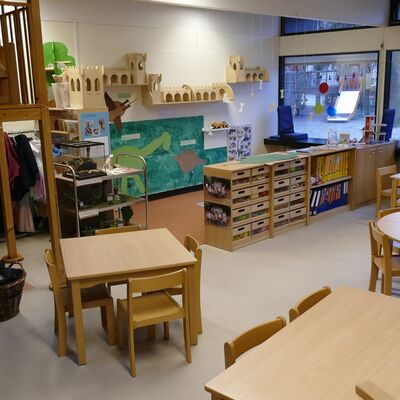 Ein Raum mit einigen Holztischen und Holzstühlen, Holzregalen, Spielzeug und einer großen Fensterfront.