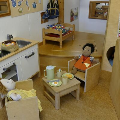 Eine kleine Ecke des Raumes mit einer kleinen Spielküche. Davor steht ein kleiner Tisch mit zwei Holzstühlen auf denen zwei Puppen mit gelbem und rotem Kleid sitzen. Im Hintergrund steht ein Puppenbett aus Holz.
