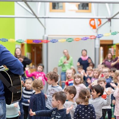 Eine Gruppe Kindergartenkinder stehen vor einem Musiker mit Gitarre und tanzen. Im Hintergrund stehen einige Erzieherinnen.
